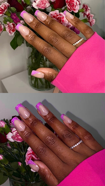 Close-up of a hand with pink reflective glitter French tip design by decode beauty against a pink floral background